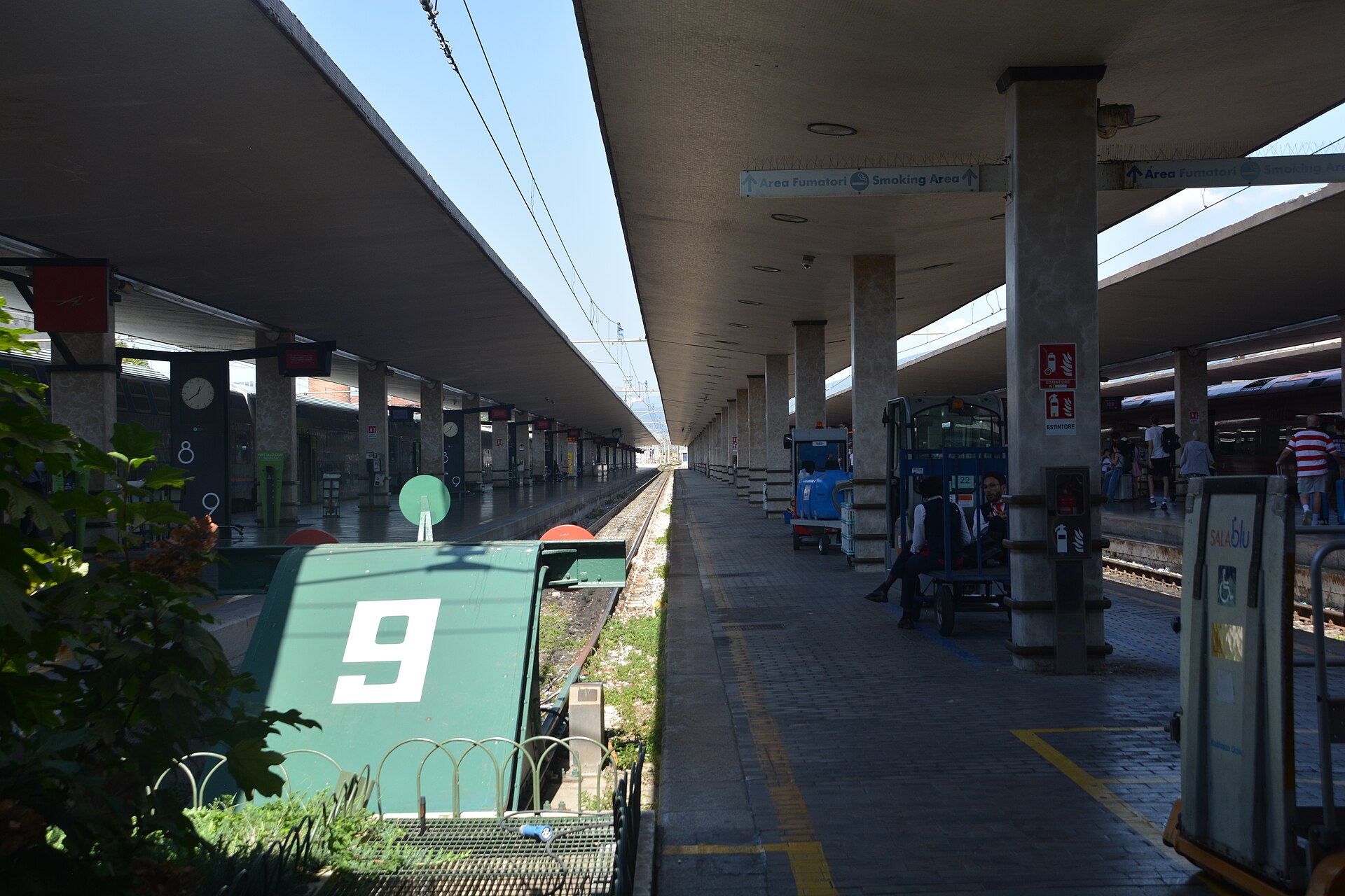 Concrete canopies shade Santa Maria Novella’s platforms in Florence, with track buffers and waiting passengers beside a train.
