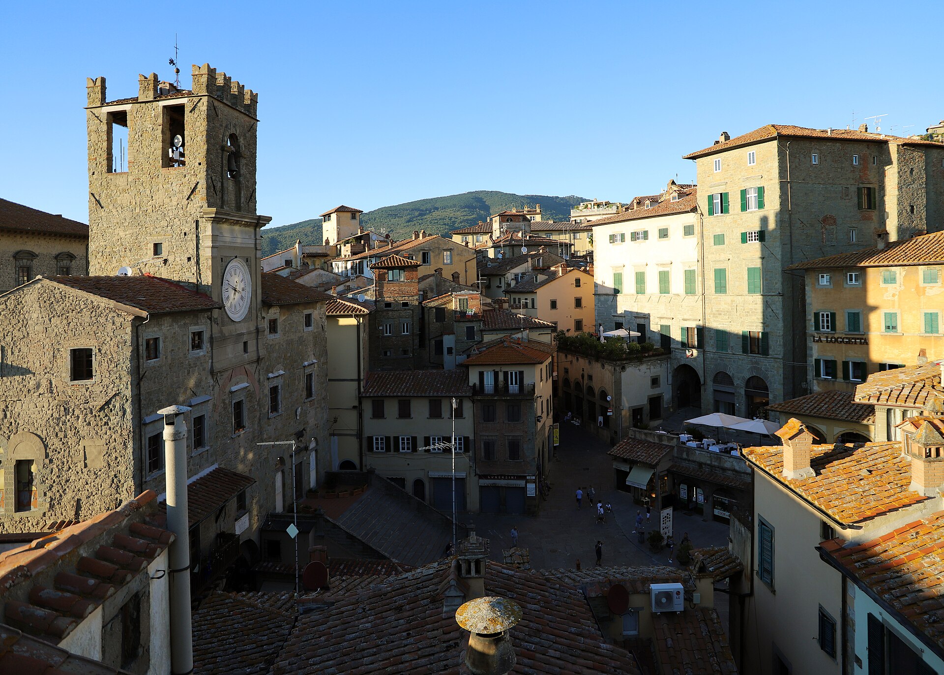 Cortona’s stone town hall and clock tower overlook Piazza della Repubblica, enclosed by buildings with green shutters.