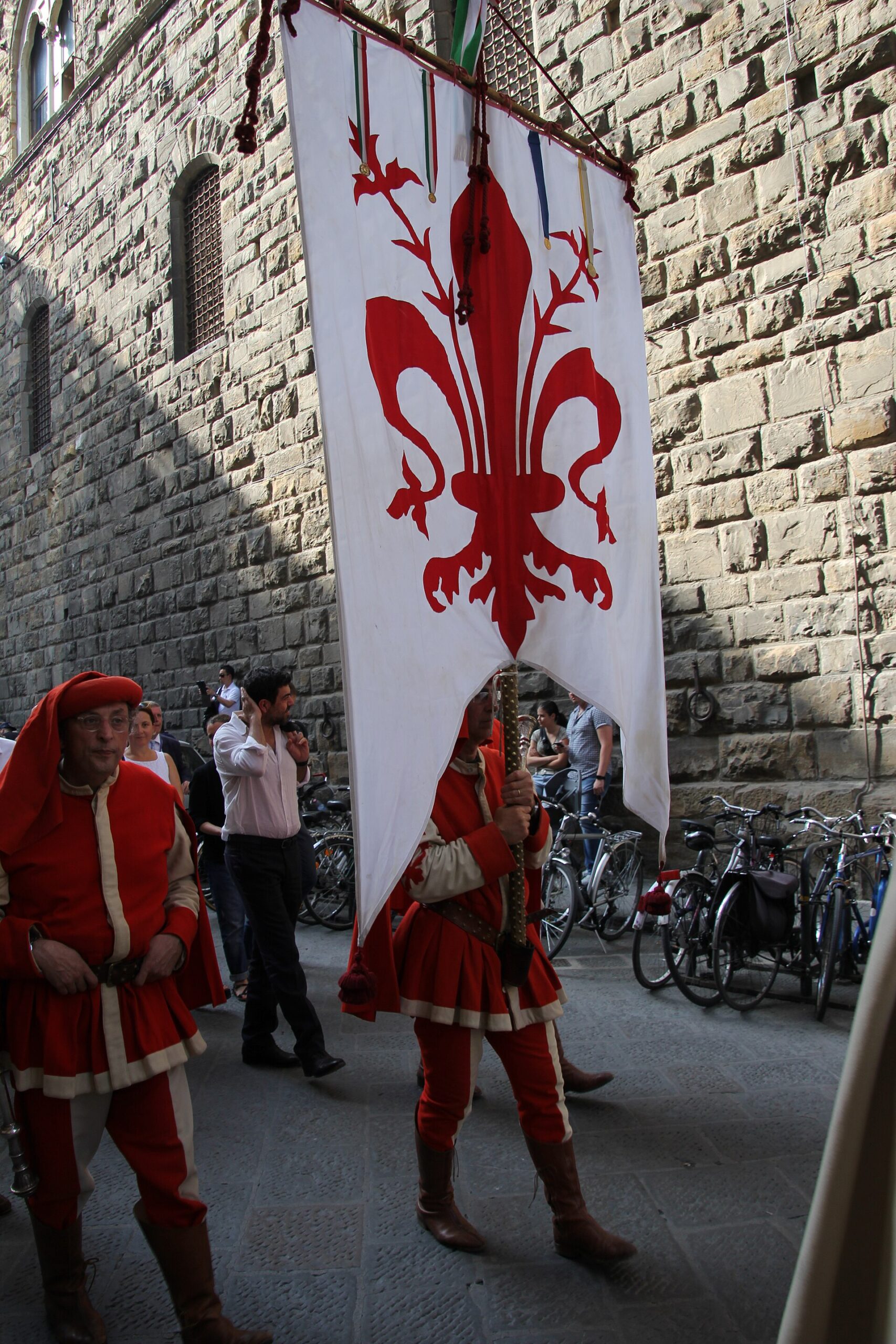 A costumed banner bearer carries Florence's red lily on white during the city's historical procession beside a stone palace wall.