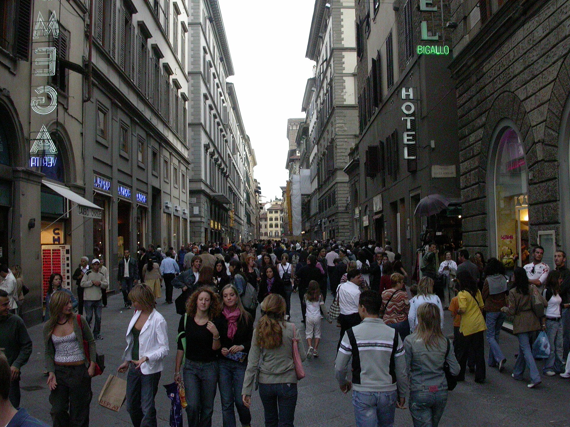 Crowds walk along Via dei Calzaiuoli in central Florence, between stone frontages and rows of shuttered windows.
