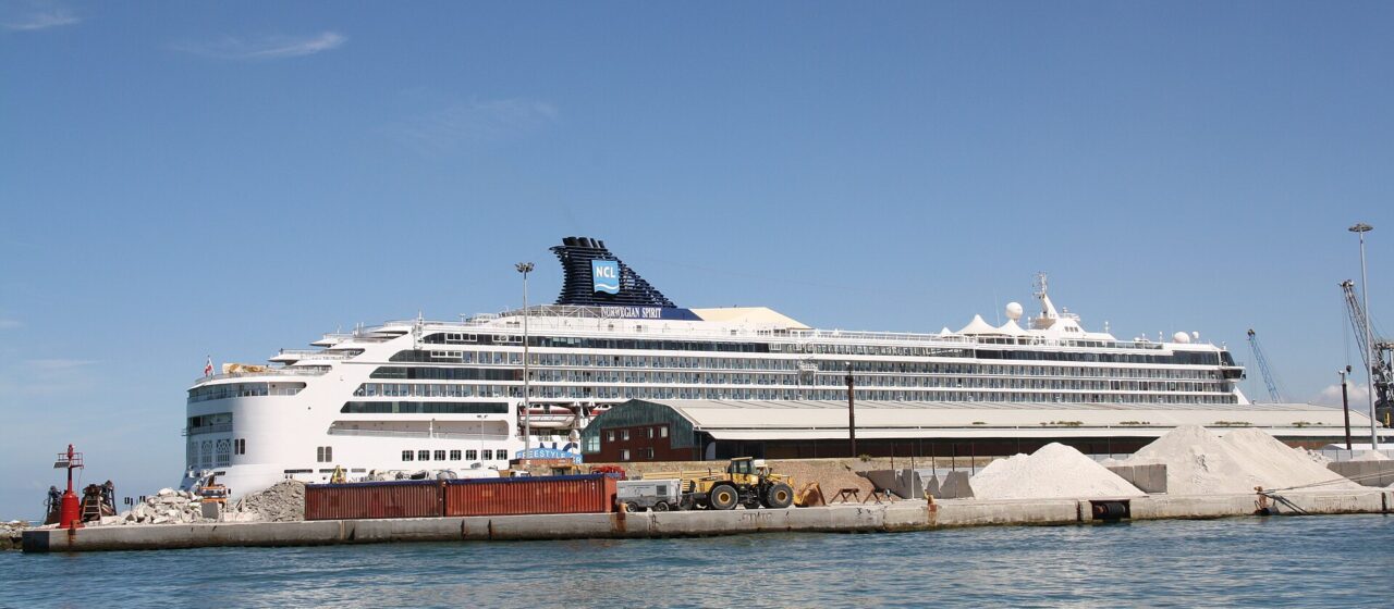 A large white cruise ship moored inside the breakwater of Livorno harbour, with a red harbour light and piles of quarried stone on the quay in front