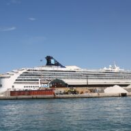 A large white cruise ship moored inside the breakwater of Livorno harbour, with a red harbour light and piles of quarried stone on the quay in front