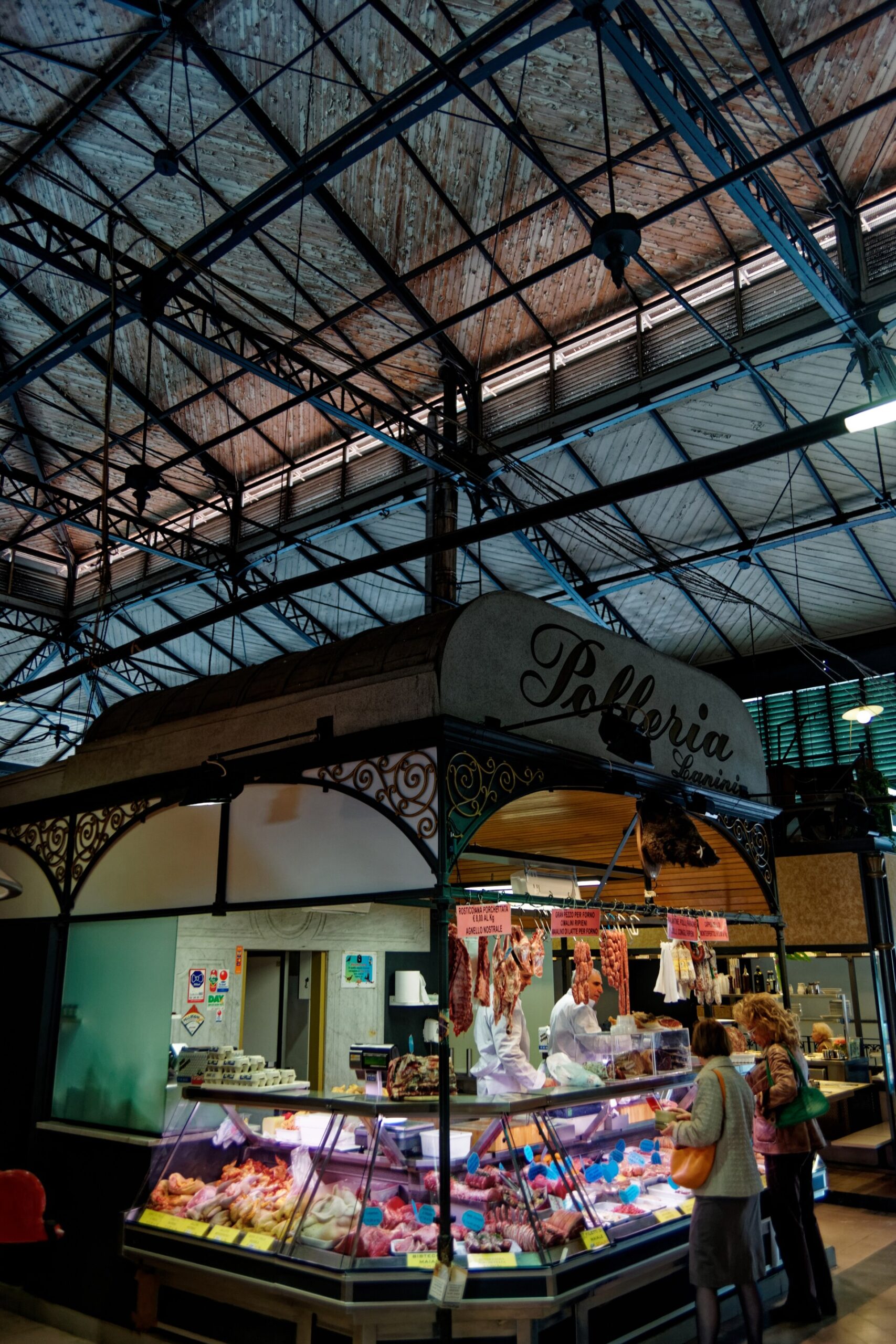 Customers browse a poultry counter beneath blue iron roof trusses inside Florence's Sant'Ambrogio market hall.