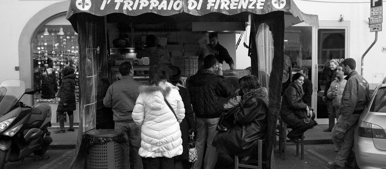 Customers gather at a lampredotto kiosk beneath the I' Trippaio di Firenze awning on Florence's Via Gioberti.