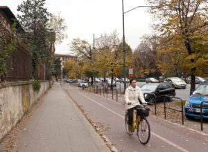 A cyclist rides a yellow bicycle along a separated Florence cycle path beside autumn trees and roadside railings.