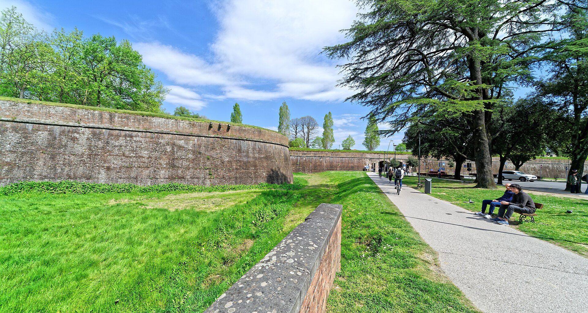 A cyclist and walkers pass Lucca’s brick ramparts beside a grassy ditch and a shaded bench.