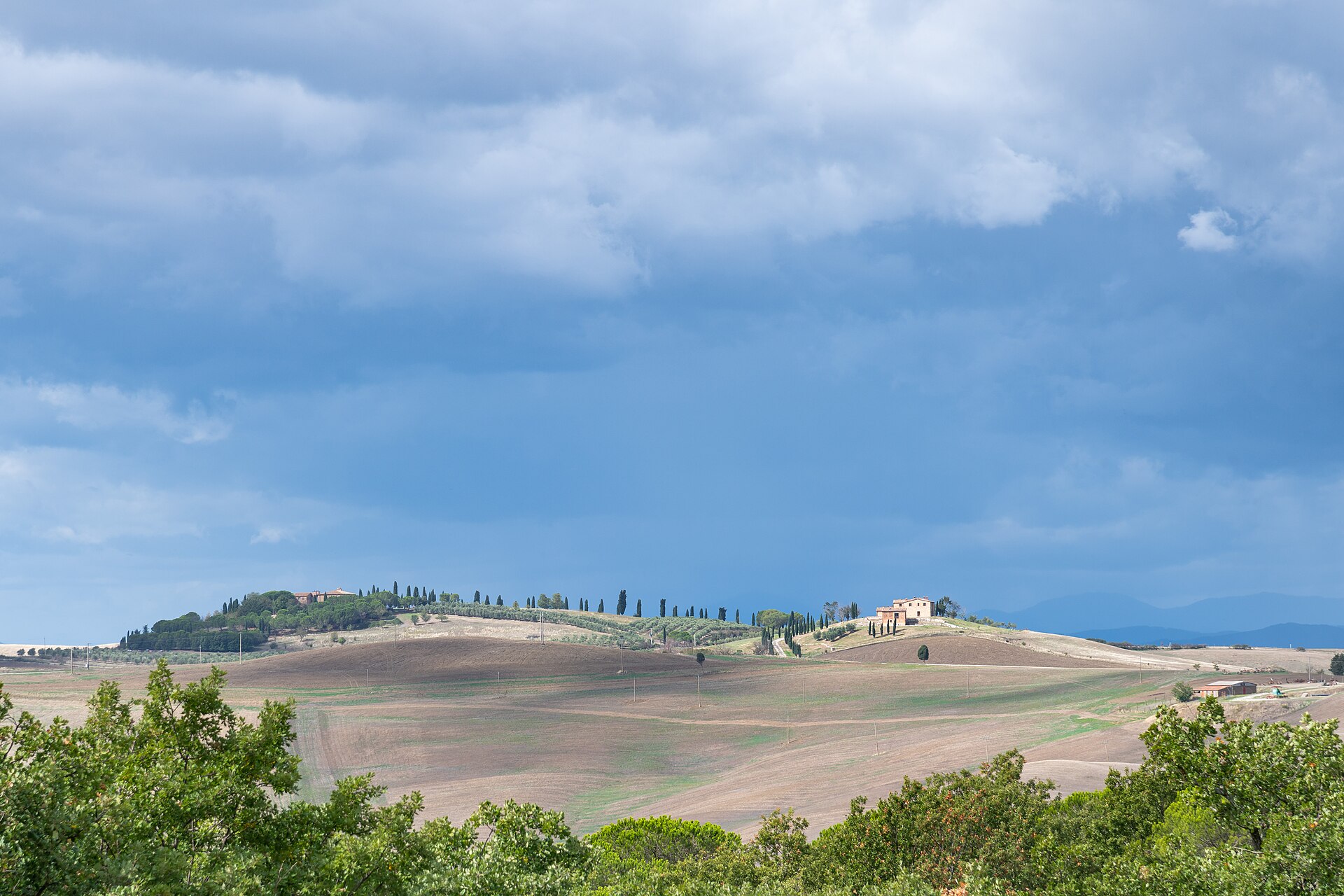 Cypress-lined ridges and farm buildings rise above rolling cultivated fields in the Val d’Orcia under gathering clouds.