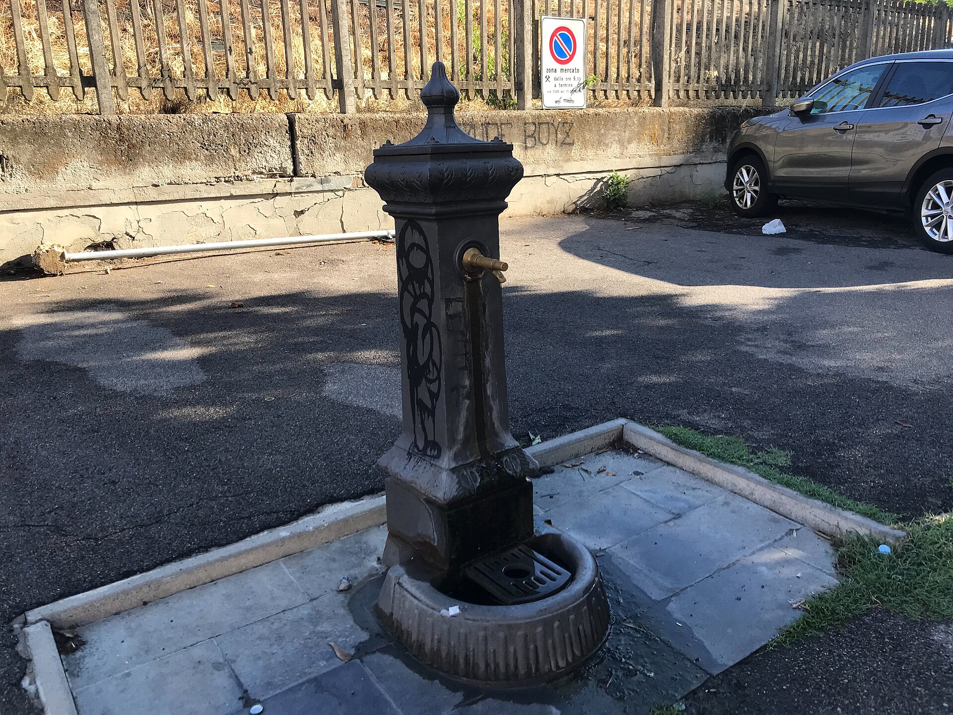 A dark cast-iron drinking fountain with a brass spout and drain basin stands beside a street in Florence.