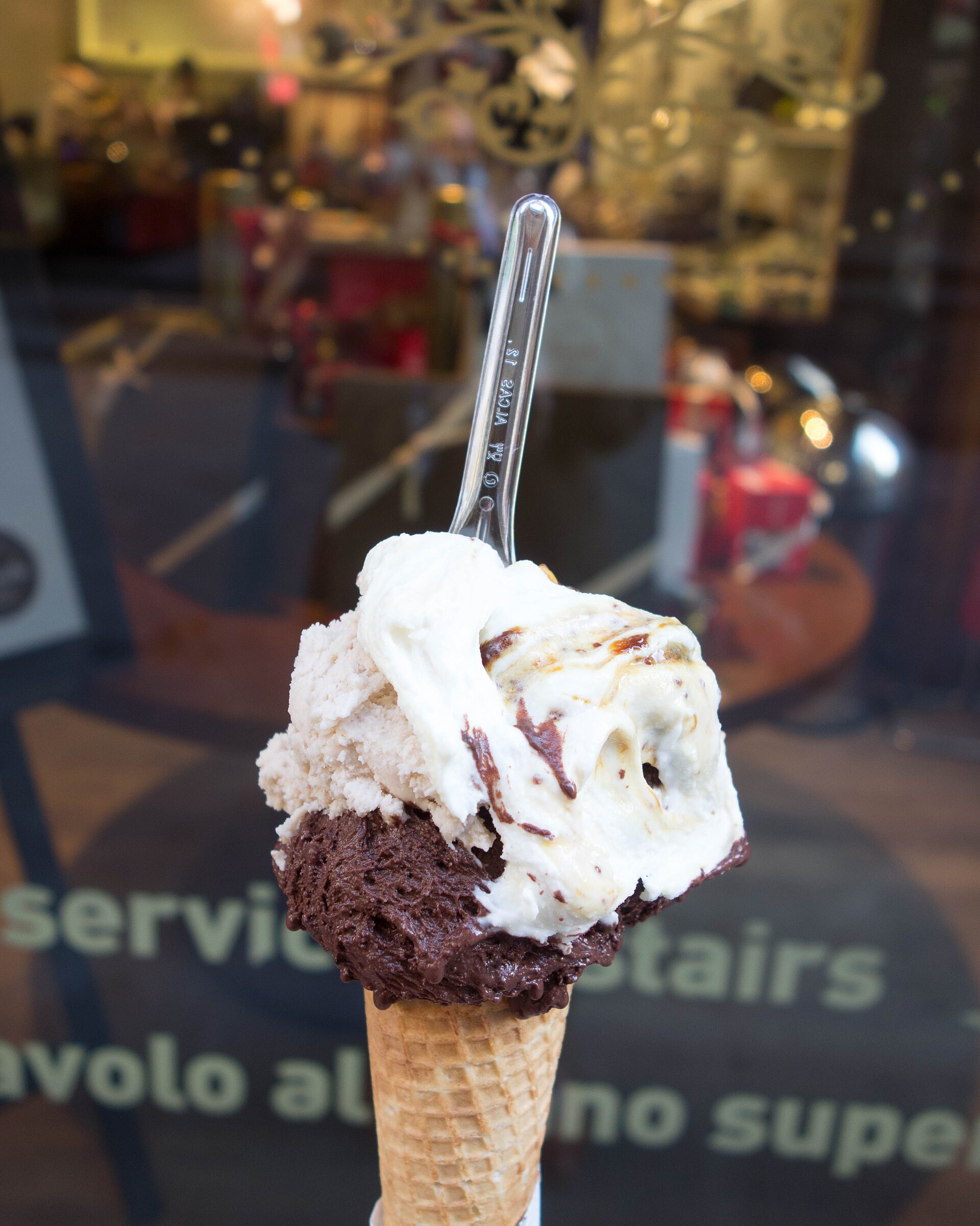 Dark and pale gelato fills a waffle cone outside a Florence shop window, with a clear spoon tucked into it.