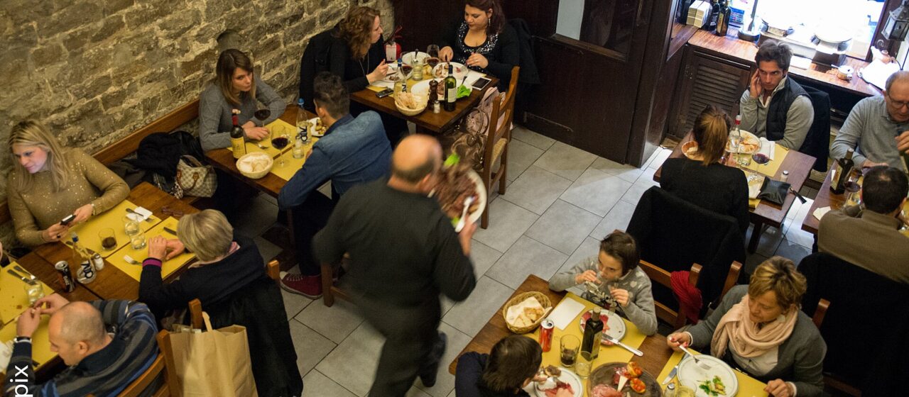 Diners share small wooden tables beside a stone wall as a waiter crosses a busy Florence osteria dining room.
