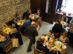 Diners share small wooden tables beside a stone wall as a waiter crosses a busy Florence osteria dining room.
