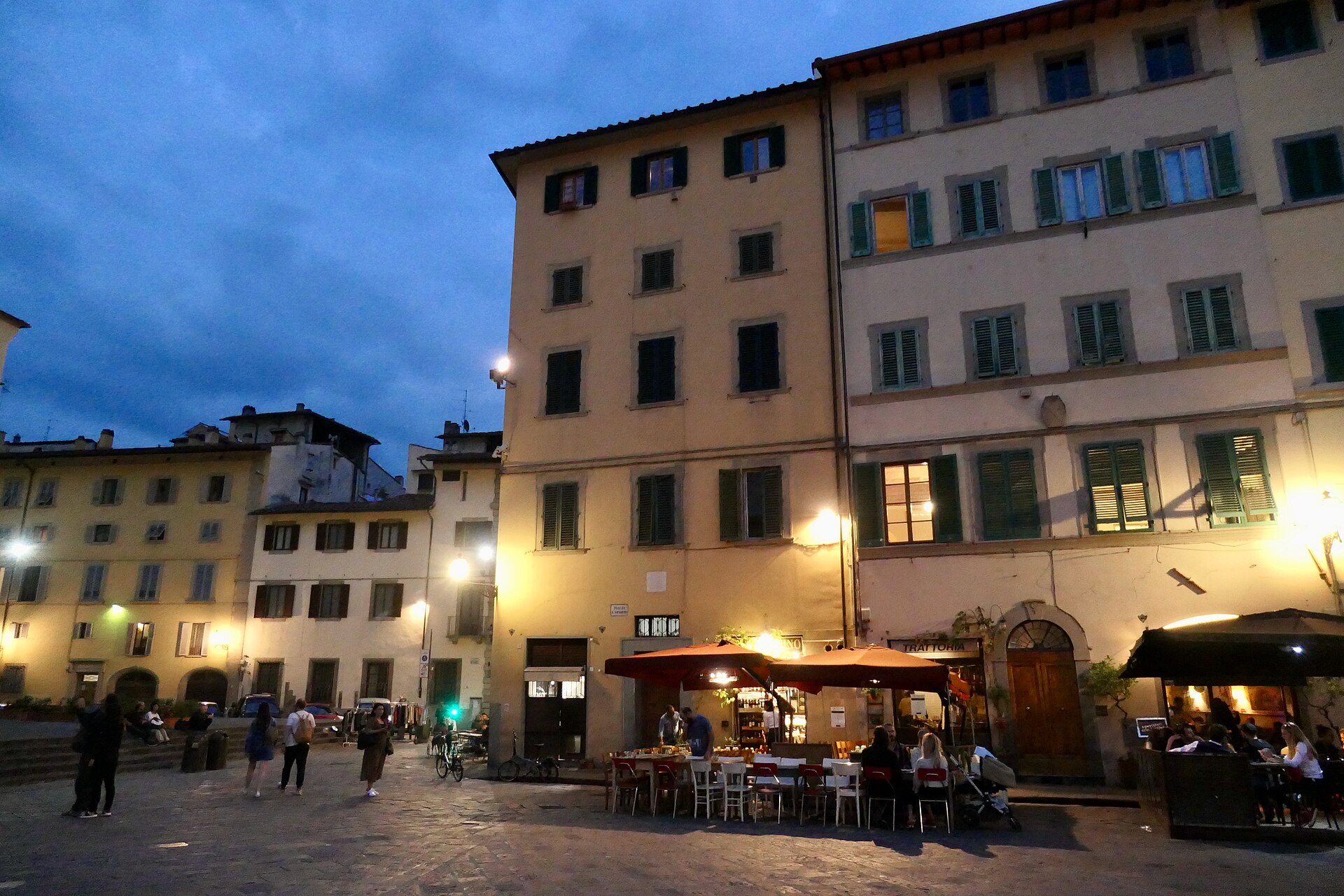 Diners sit beneath umbrellas as lamps illuminate the buildings lining Piazza Santo Spirito in Florence at dusk.