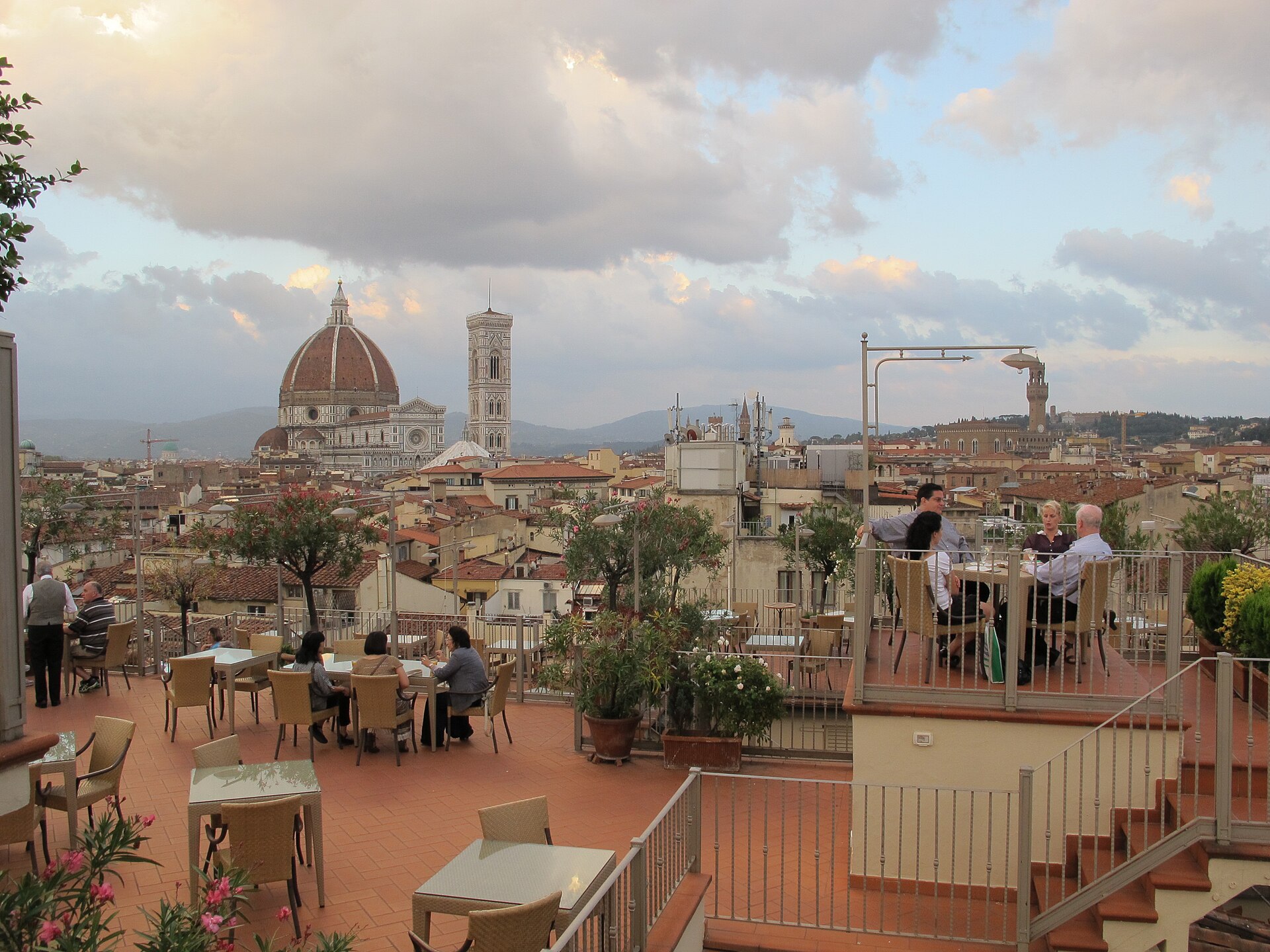 Diners at tables on a hotel rooftop terrace at dusk, with the Florence Duomo dome and Giotto's bell tower rising above the terracotta roofs