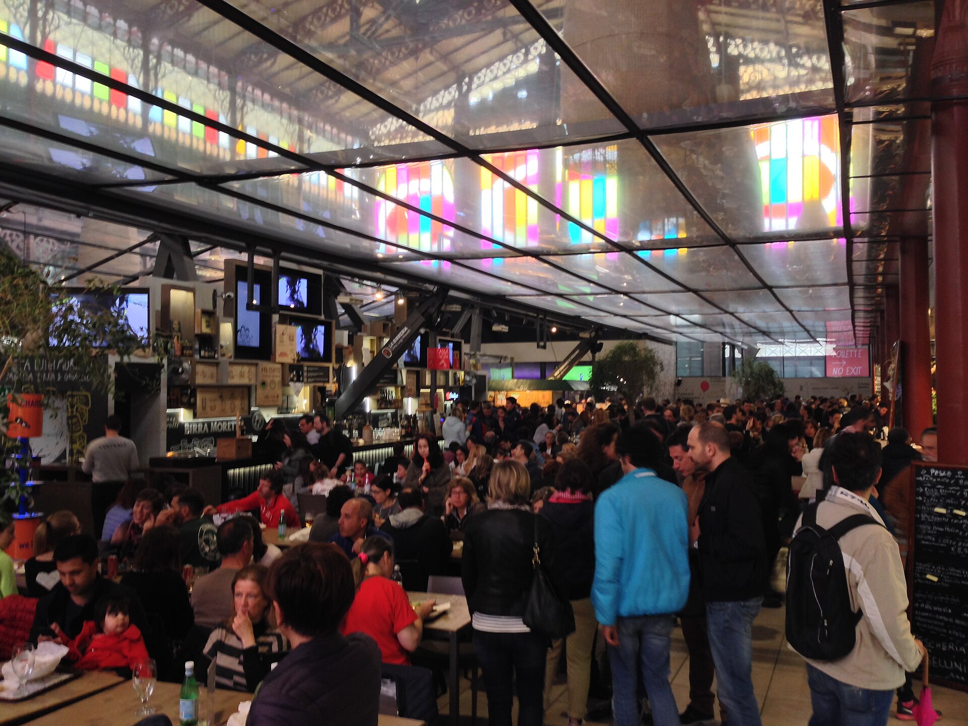 Diners and visitors crowd communal tables and food counters beneath a reflective canopy in Florence's Mercato Centrale upstairs food hall.