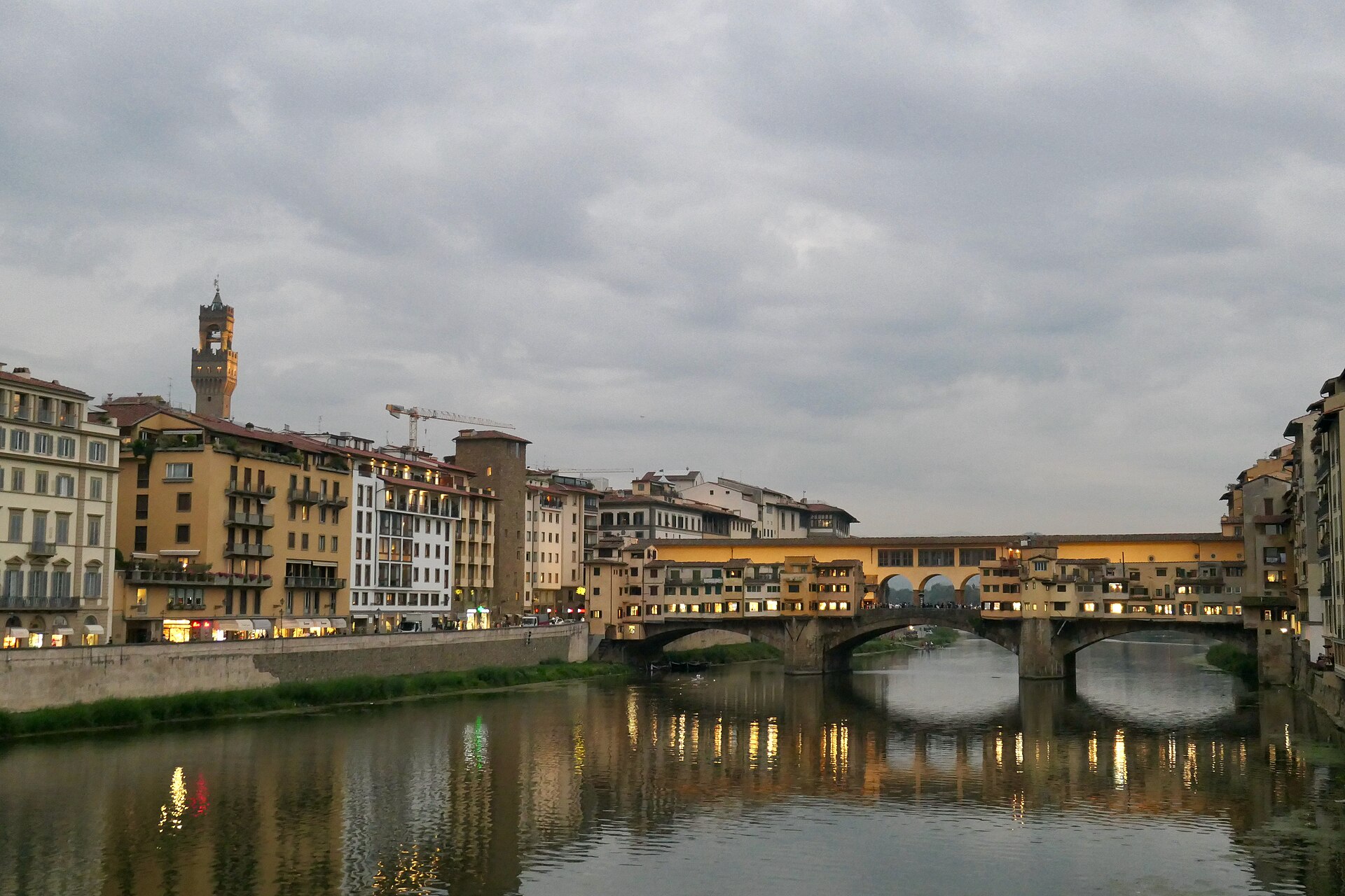 Evening lights reflect beneath Ponte Vecchio in Florence, with Palazzo Vecchio’s tower rising above the riverfront under grey clouds.