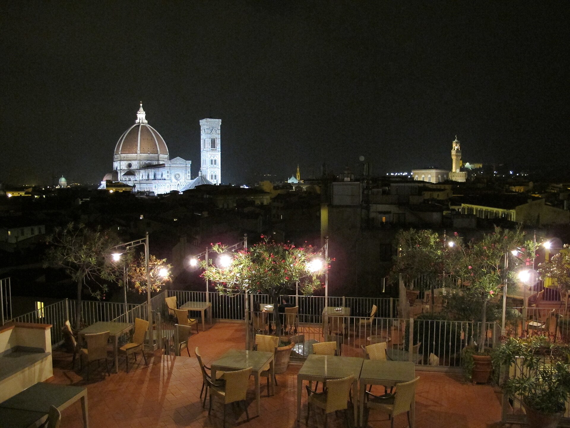 Floodlit Florence Duomo and bell tower at night seen across a rooftop terrace with lamps, tables and wicker chairs