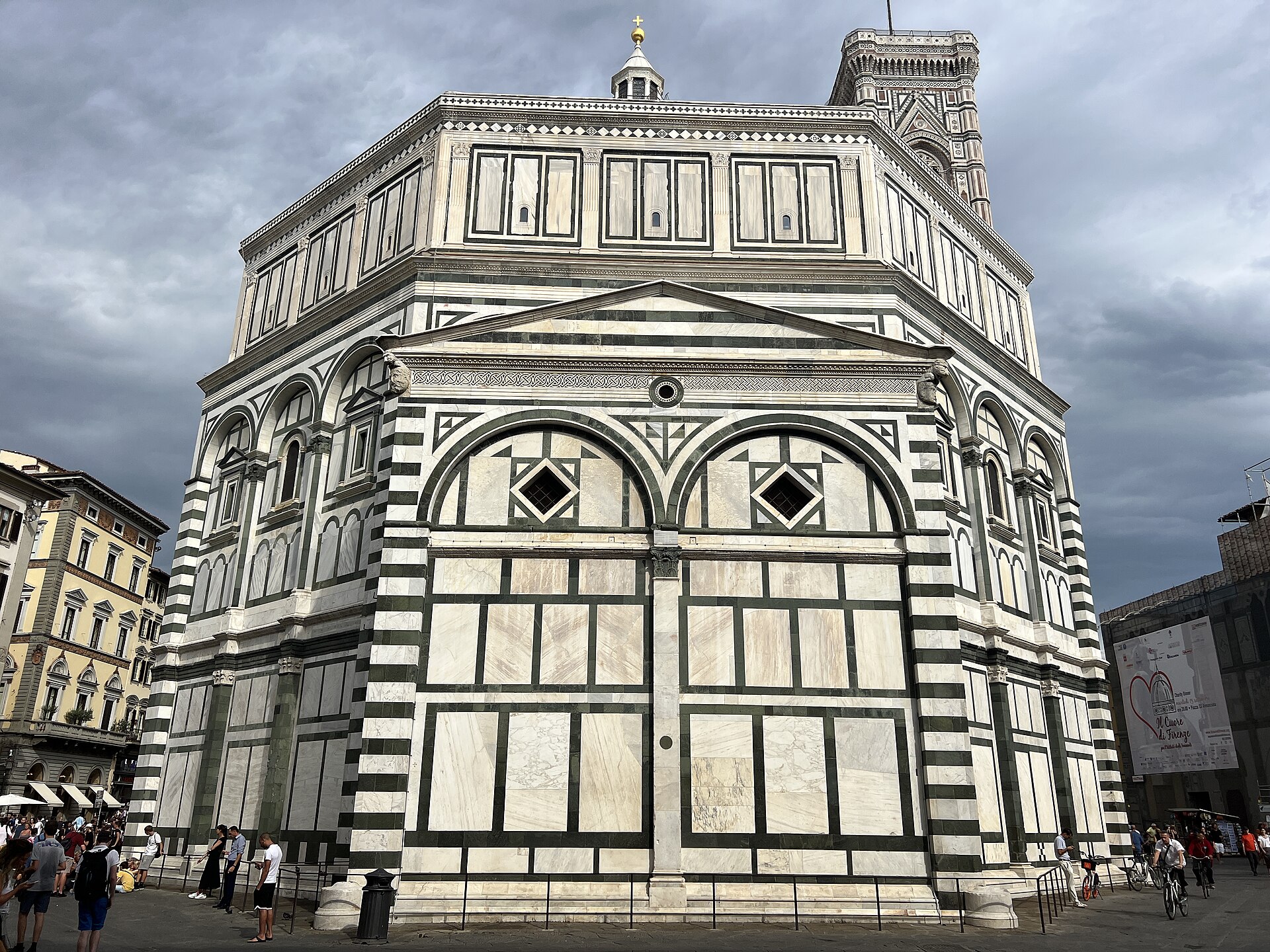 The Florence Baptistery's projecting apse and octagonal upper walls show geometric green and white marble beneath a cloudy sky, with visitors below.