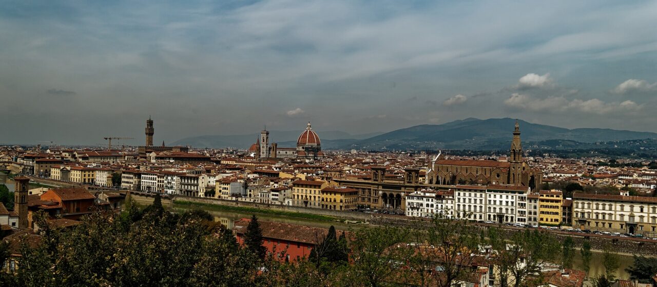 Florence rooftops stretch along the Arno beneath Brunelleschi's dome, Giotto's bell tower and the hills under a cloudy sky.
