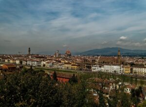Florence rooftops stretch along the Arno beneath Brunelleschi's dome, Giotto's bell tower and the hills under a cloudy sky.