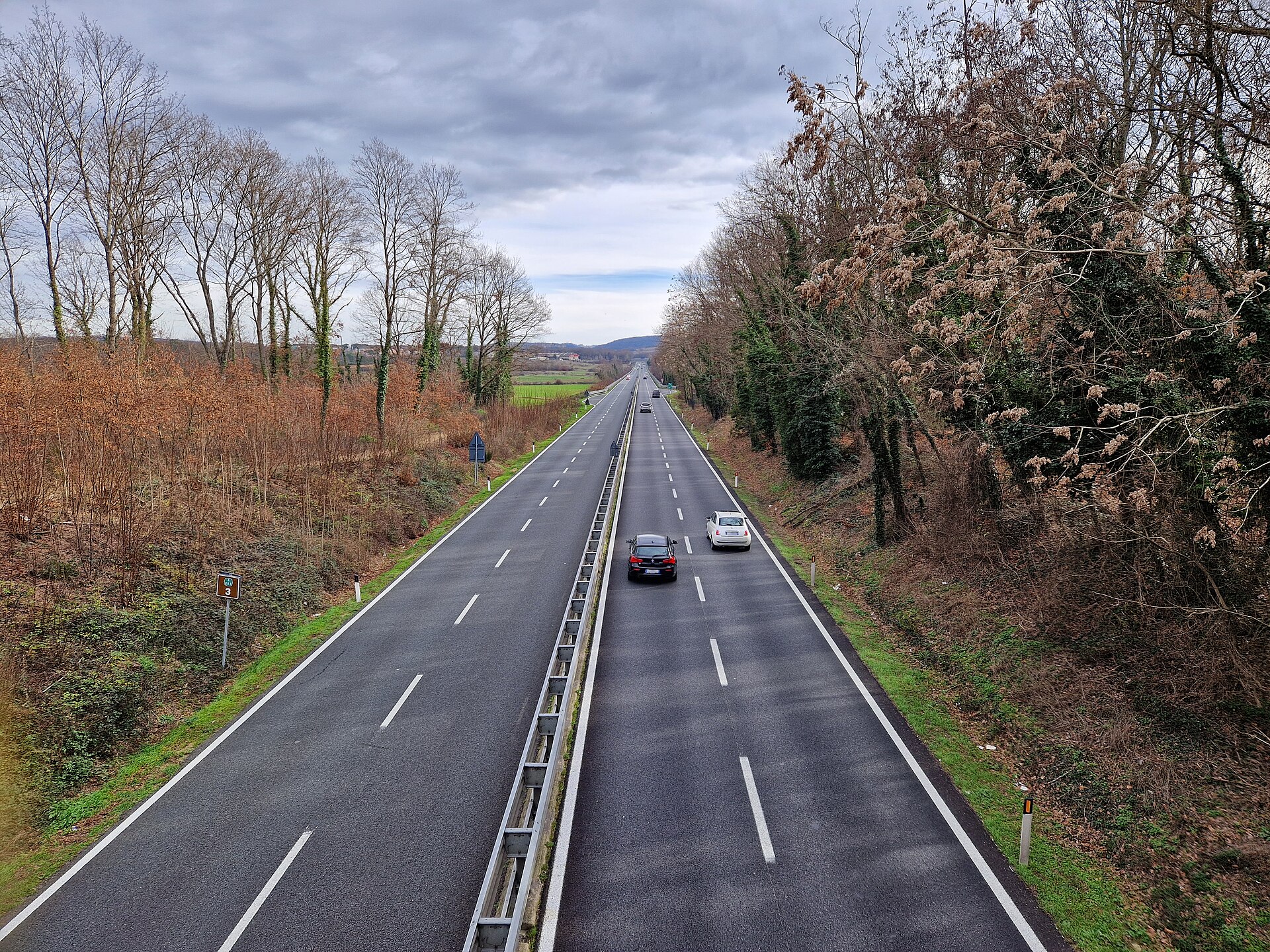 A straight four-lane dual carriageway running through Tuscan countryside between rows of bare winter trees, with a couple of cars in the distance