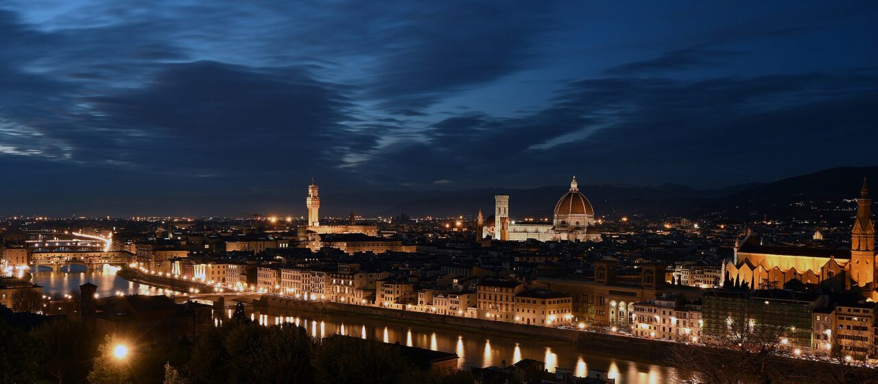 Florence's cathedral dome and Palazzo Vecchio rise above illuminated riverside buildings, with warm lights reflected across the Arno at night.