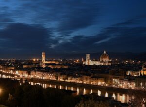 Florence's cathedral dome and Palazzo Vecchio rise above illuminated riverside buildings, with warm lights reflected across the Arno at night.