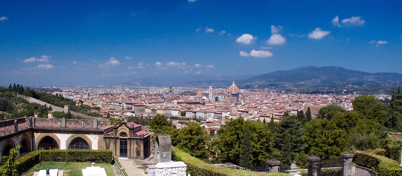 Florence's cathedral dome rises beyond green trees and the stone tombs of the San Miniato al Monte cemetery under blue skies.