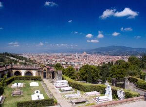 Florence's cathedral dome rises beyond green trees and the stone tombs of the San Miniato al Monte cemetery under blue skies.