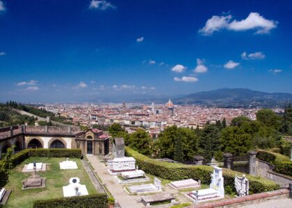 Florence's cathedral dome rises beyond green trees and the stone tombs of the San Miniato al Monte cemetery under blue skies.