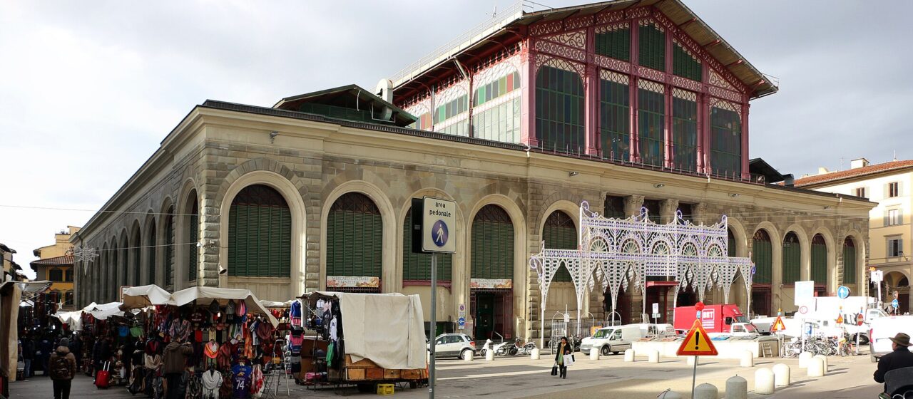 Florence's Mercato Centrale rises behind outdoor stalls and pedestrians, with arched shutters below its red iron-and-glass upper facade.