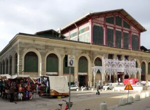 Florence's Mercato Centrale rises behind outdoor stalls and pedestrians, with arched shutters below its red iron-and-glass upper facade.