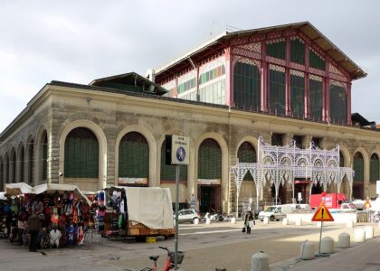 Florence's Mercato Centrale rises behind outdoor stalls and pedestrians, with arched shutters below its red iron-and-glass upper facade.