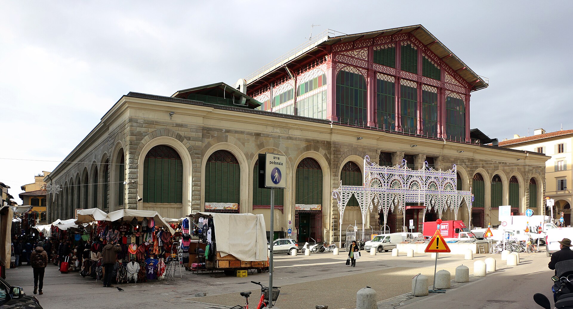 Florence's Mercato Centrale rises behind outdoor stalls and pedestrians, with arched shutters below its red iron-and-glass upper facade.