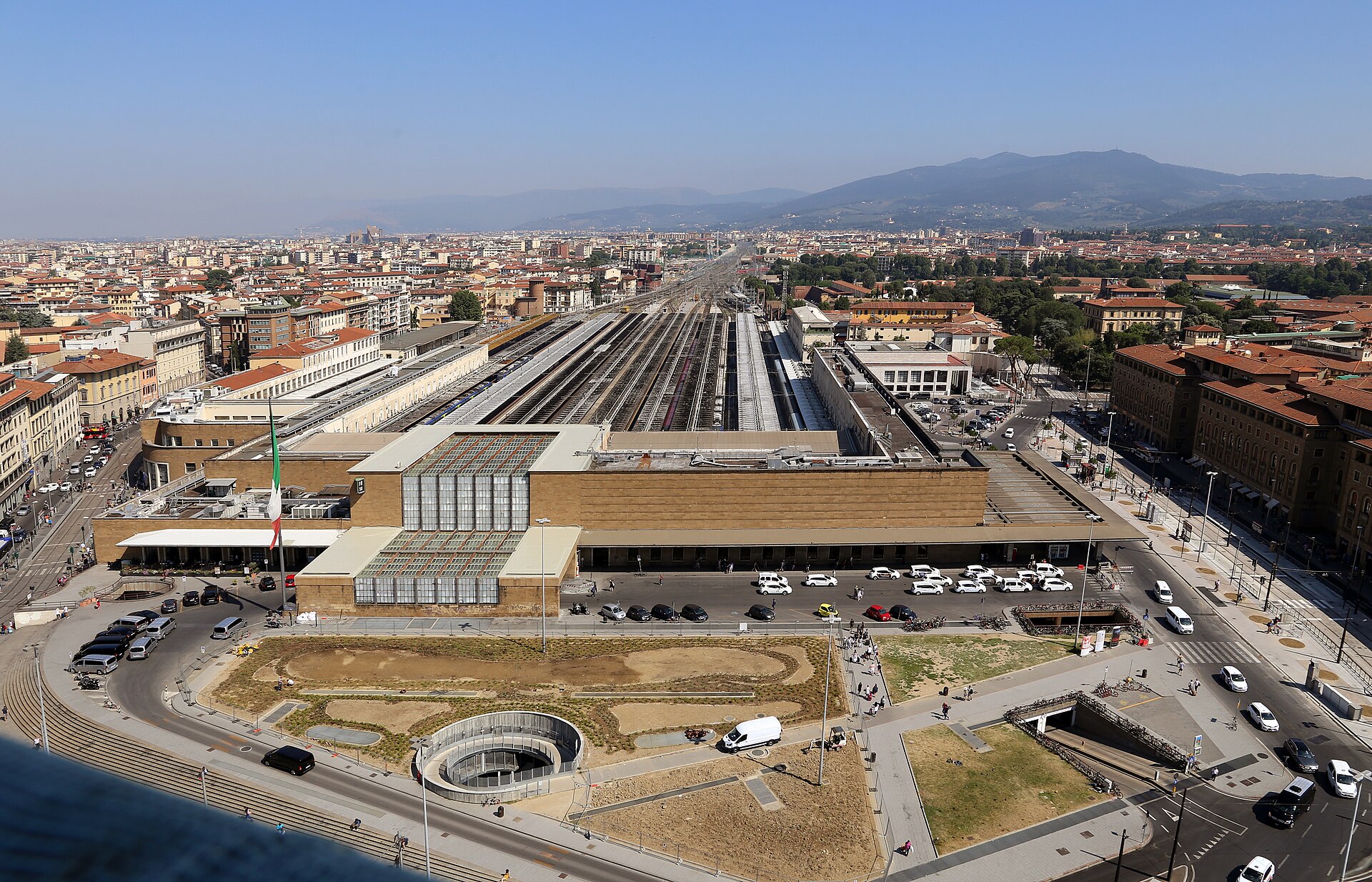 Florence’s Santa Maria Novella terminal stands below long rows of platforms, surrounded by city buildings and distant hills.