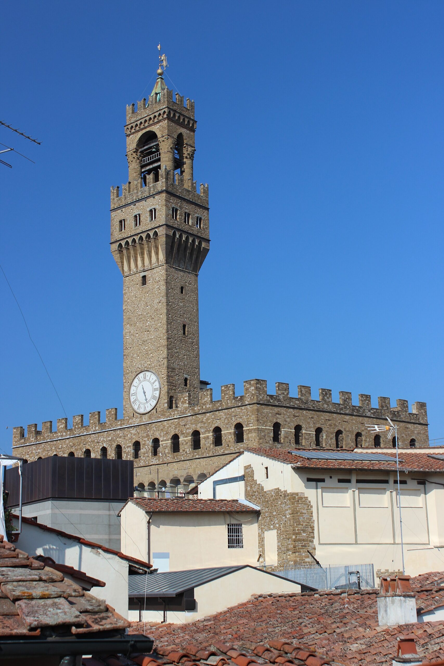 Florence's Torre di Arnolfo rises above Palazzo Vecchio's crenellated roof and nearby terracotta rooftops beneath a clear blue sky.