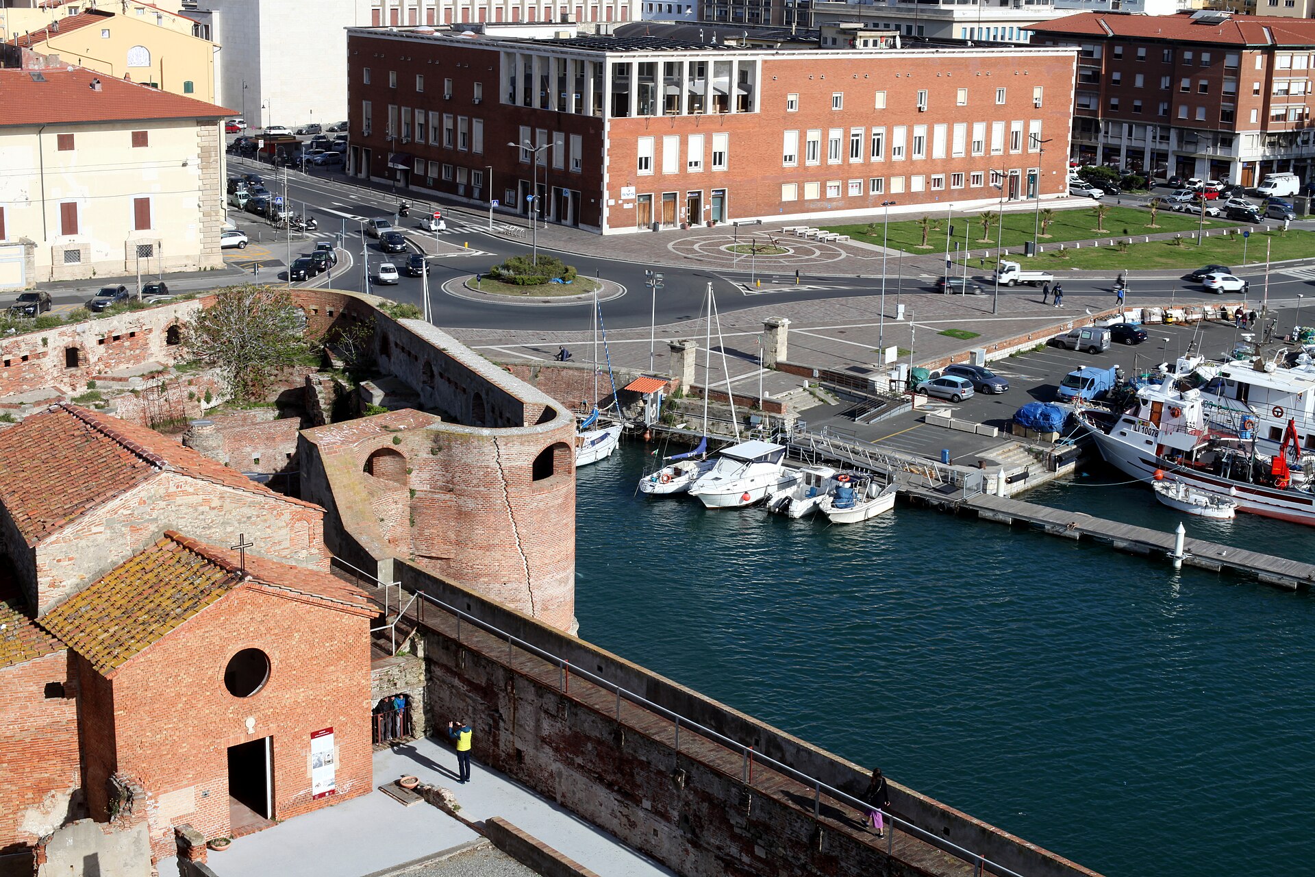 Looking down from the brick walls of the Fortezza Vecchia over Livorno's old harbour basin, with small boats moored and a busy road and modern office block behind