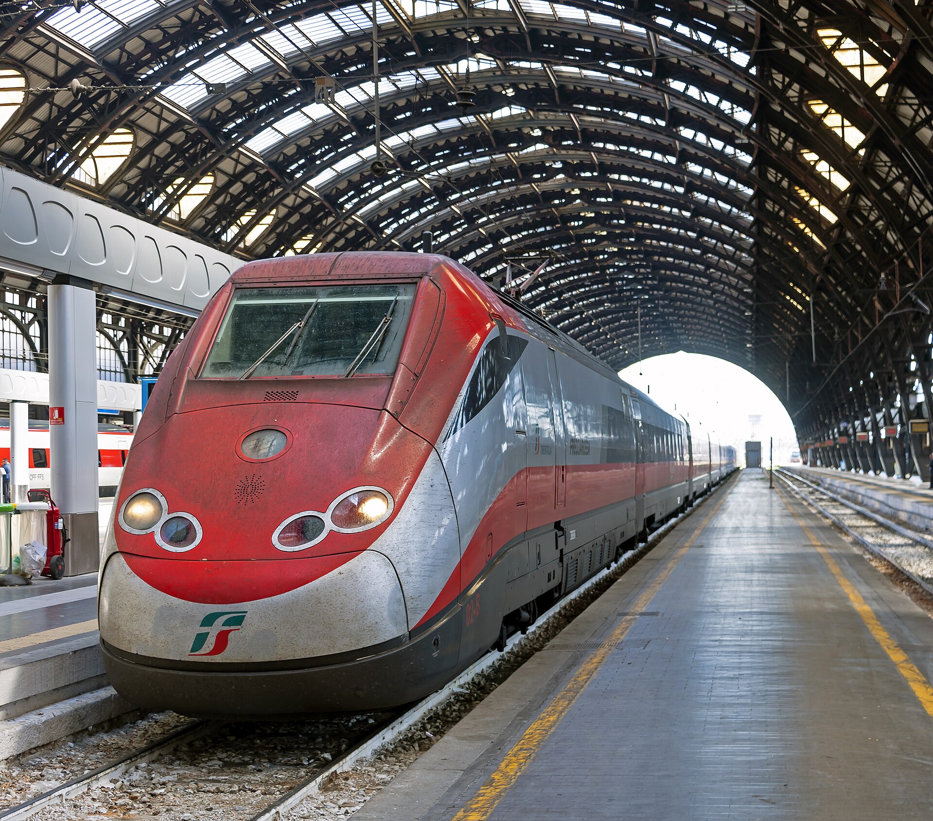 A red and silver Frecciarossa train standing at a platform under the huge arched iron and glass train shed of Milano Centrale