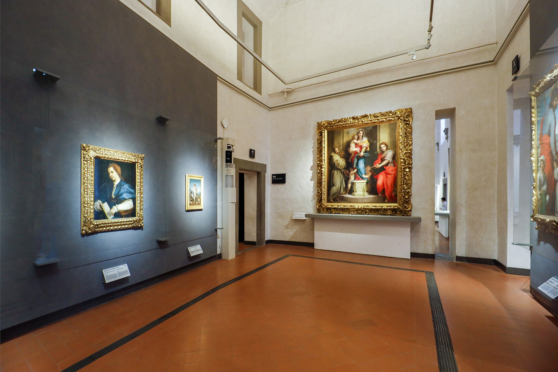 Gilded Renaissance paintings hang on grey walls around a terracotta floor in the Uffizi Gallery's Andrea del Sarto room.