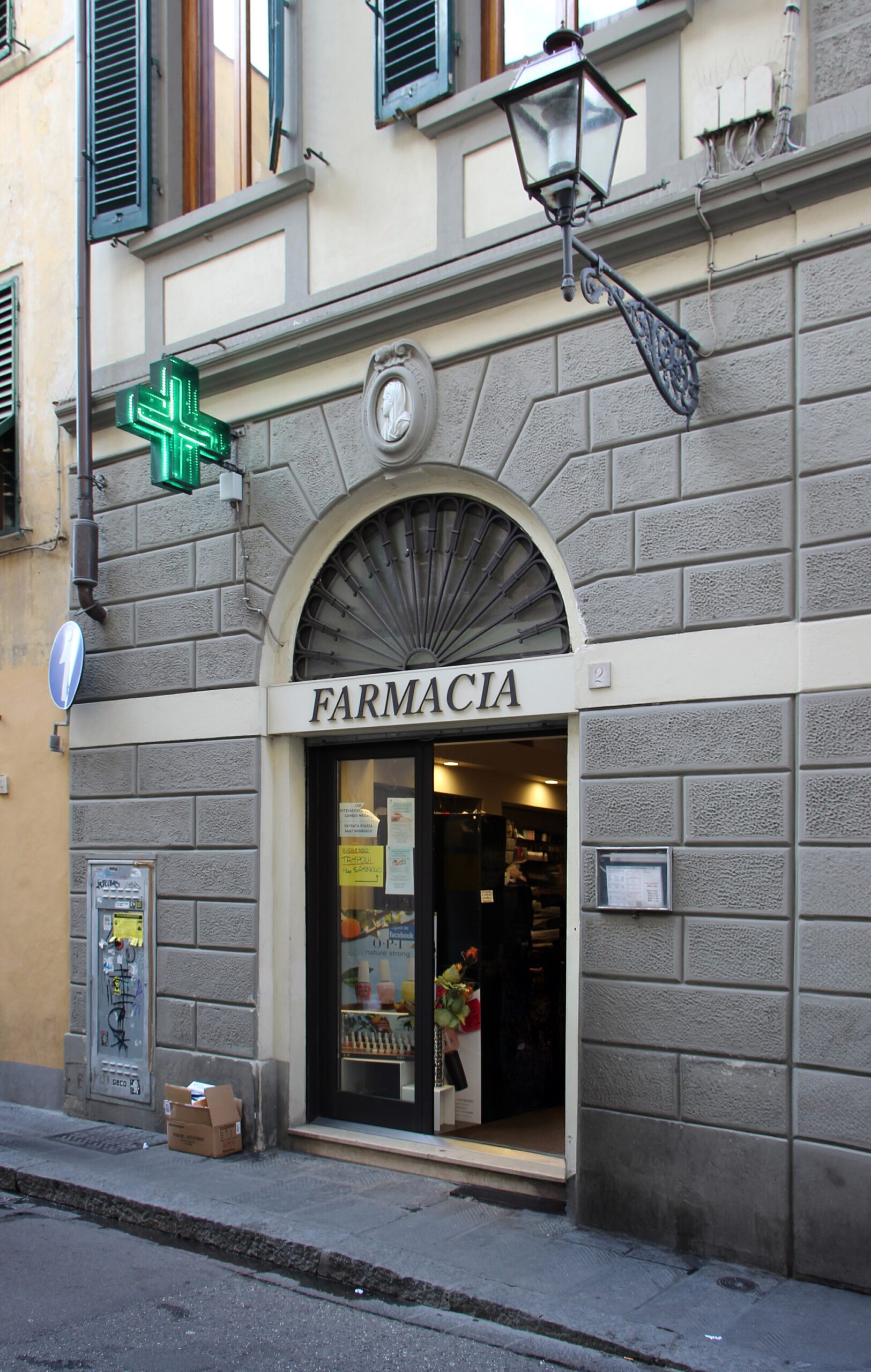 A glowing green cross hangs beside the open pharmacy doorway on Florence's Via Pietrapiana, beneath an iron street lantern.
