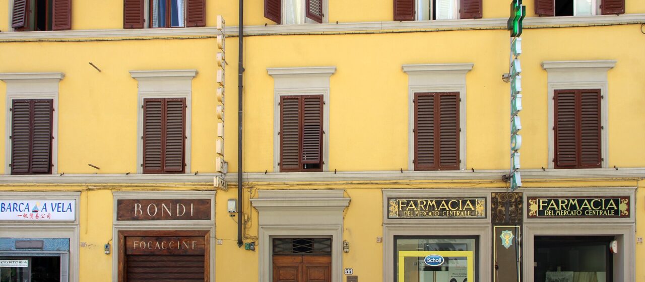 A green cross and black-and-gold pharmacy signs mark the yellow frontage of Florence's Farmacia del Mercato Centrale.