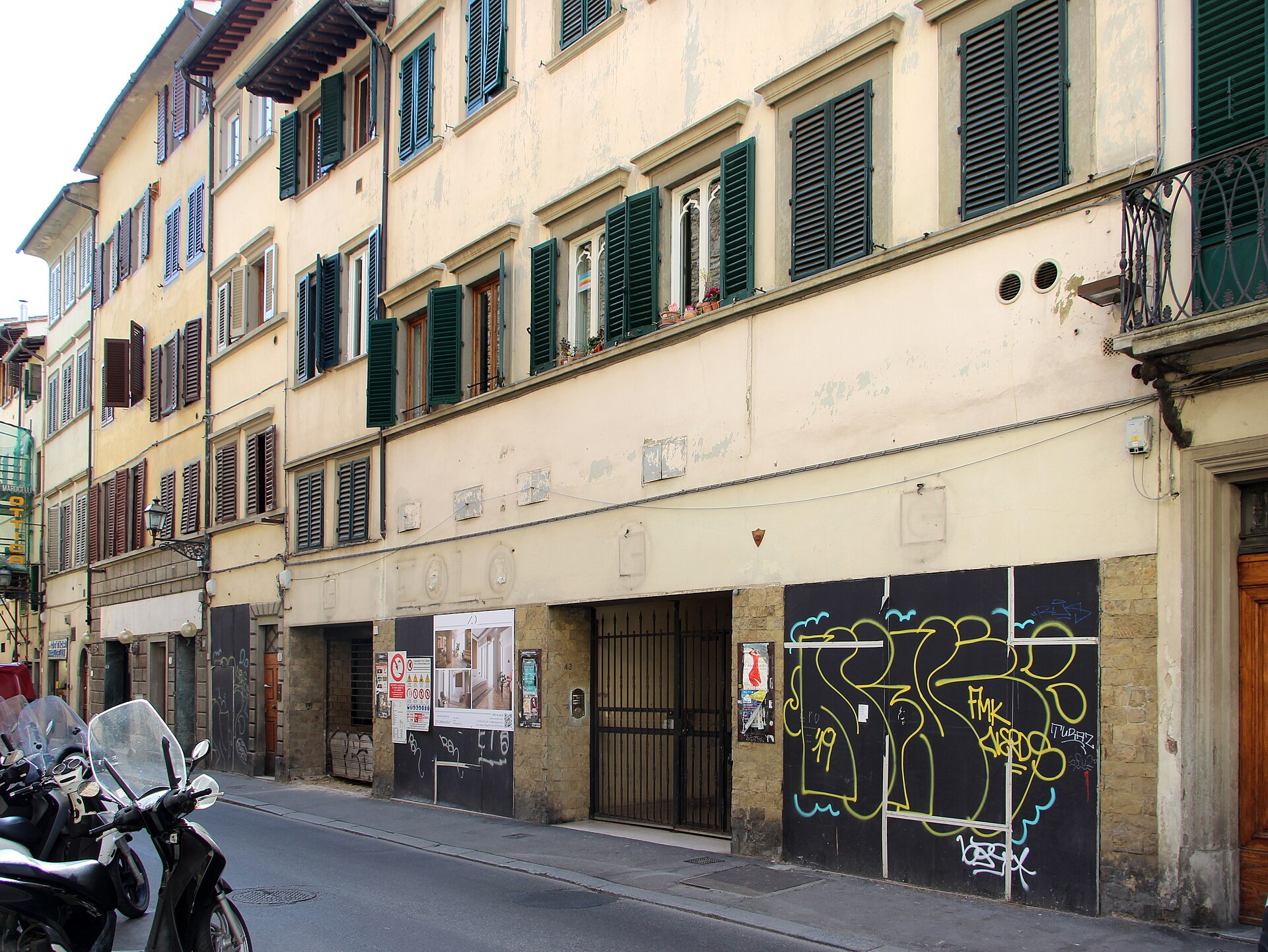 Green shutters and pale facades line Borgo San Frediano in Florence, above closed storefronts beside a narrow roadway.