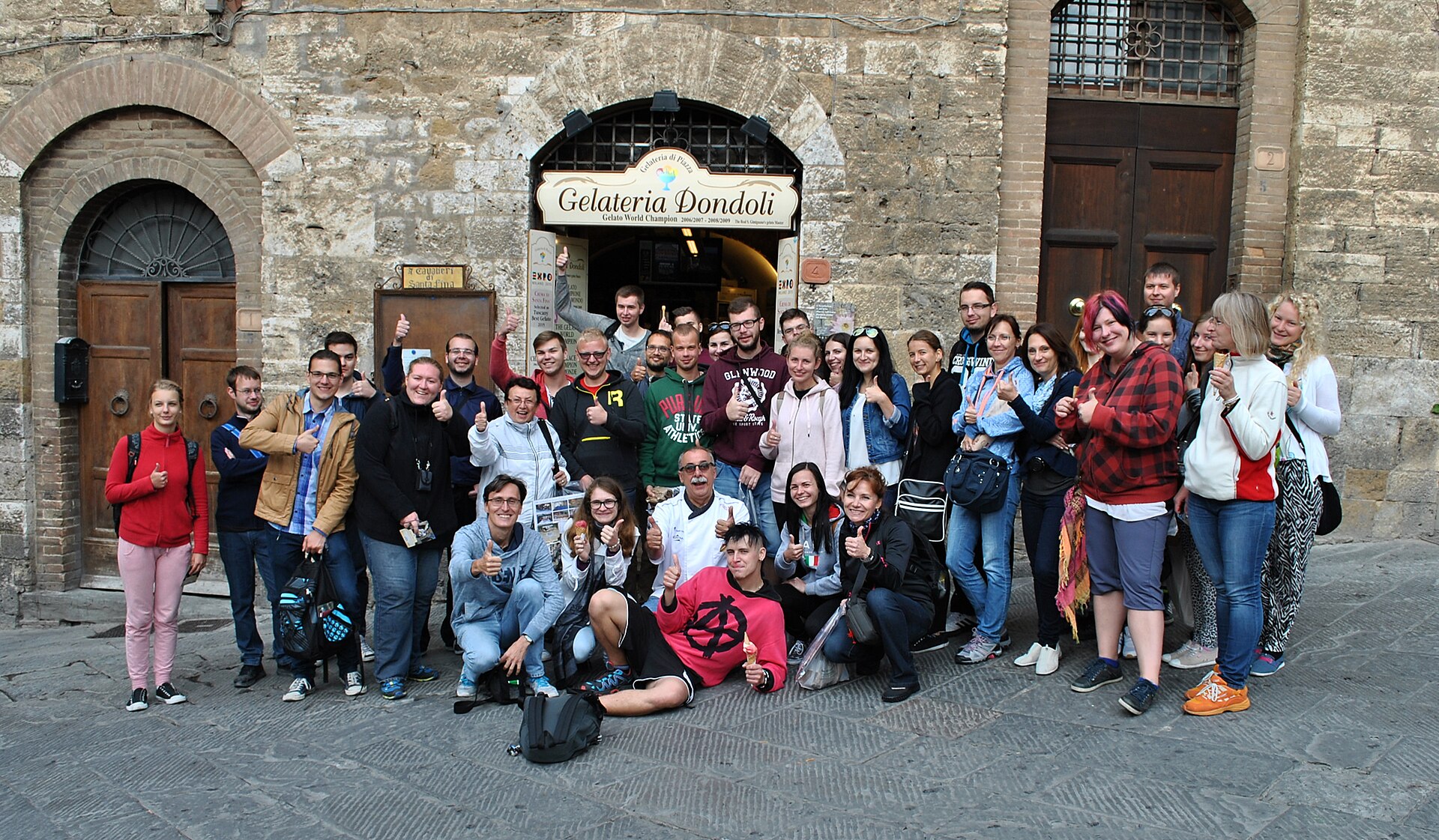 A group holding gelato gathers beneath the Gelateria Dondoli sign outside its stone shopfront in San Gimignano.