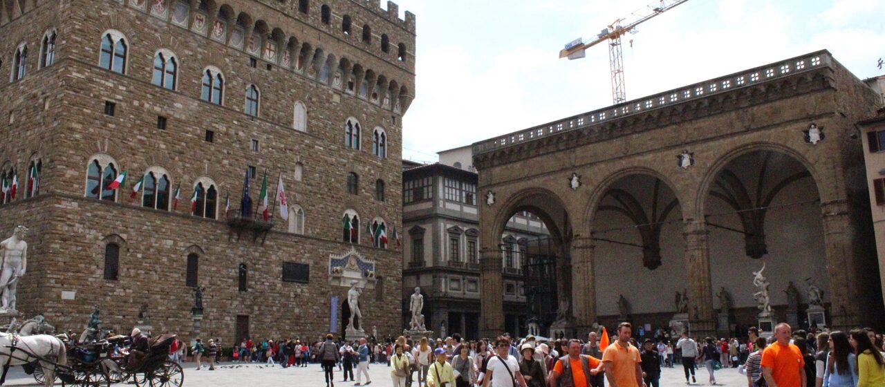 Groups of visitors cross Florence’s Piazza della Signoria between Palazzo Vecchio and the open arches of the Loggia dei Lanzi.