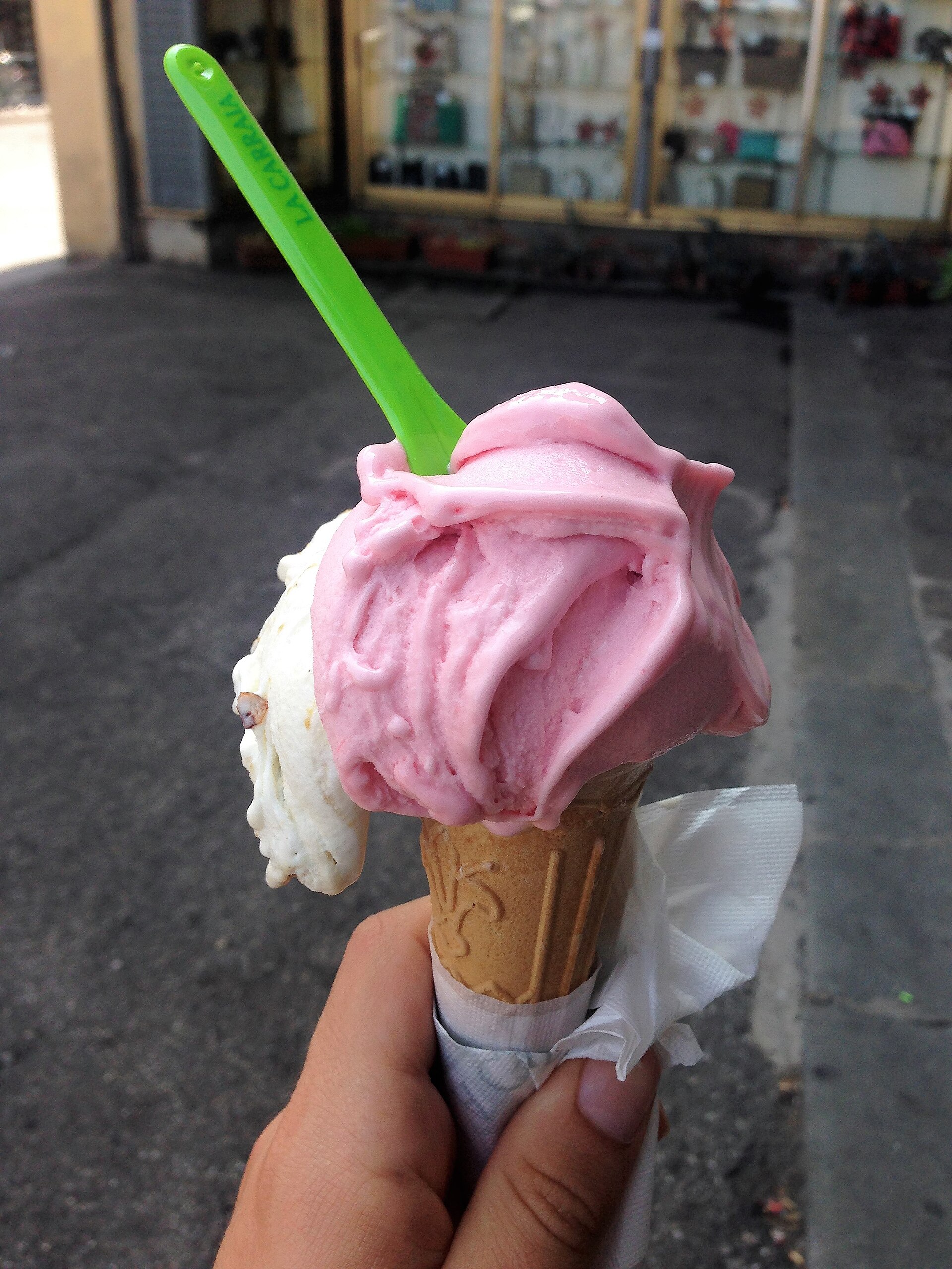 A hand holds a pink and white gelato cone with a green spoon on a paved Florence street.