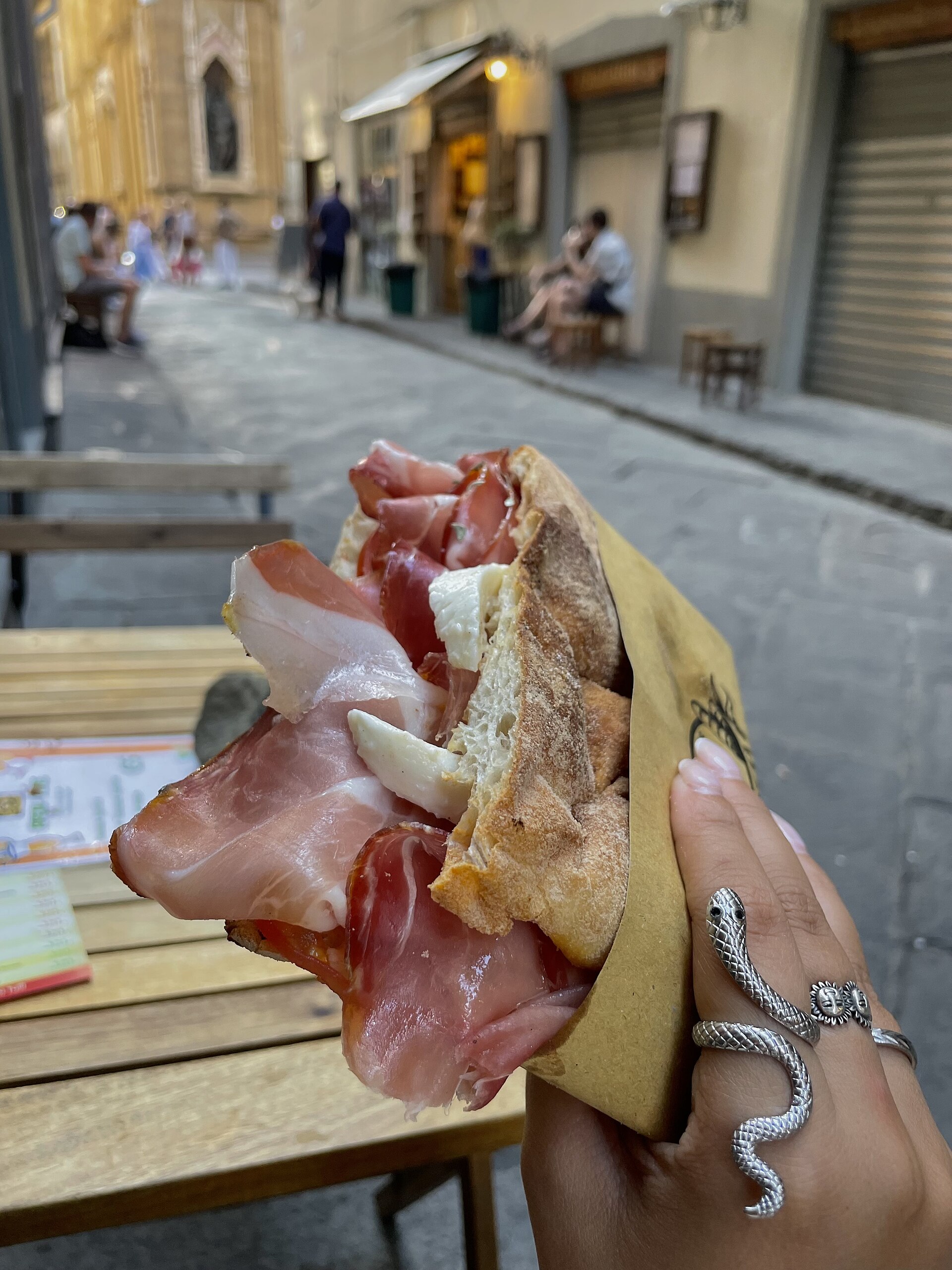 A hand holds a schiacciata sandwich filled with cured meat and pale cheese beside a stone-paved street in central Florence.