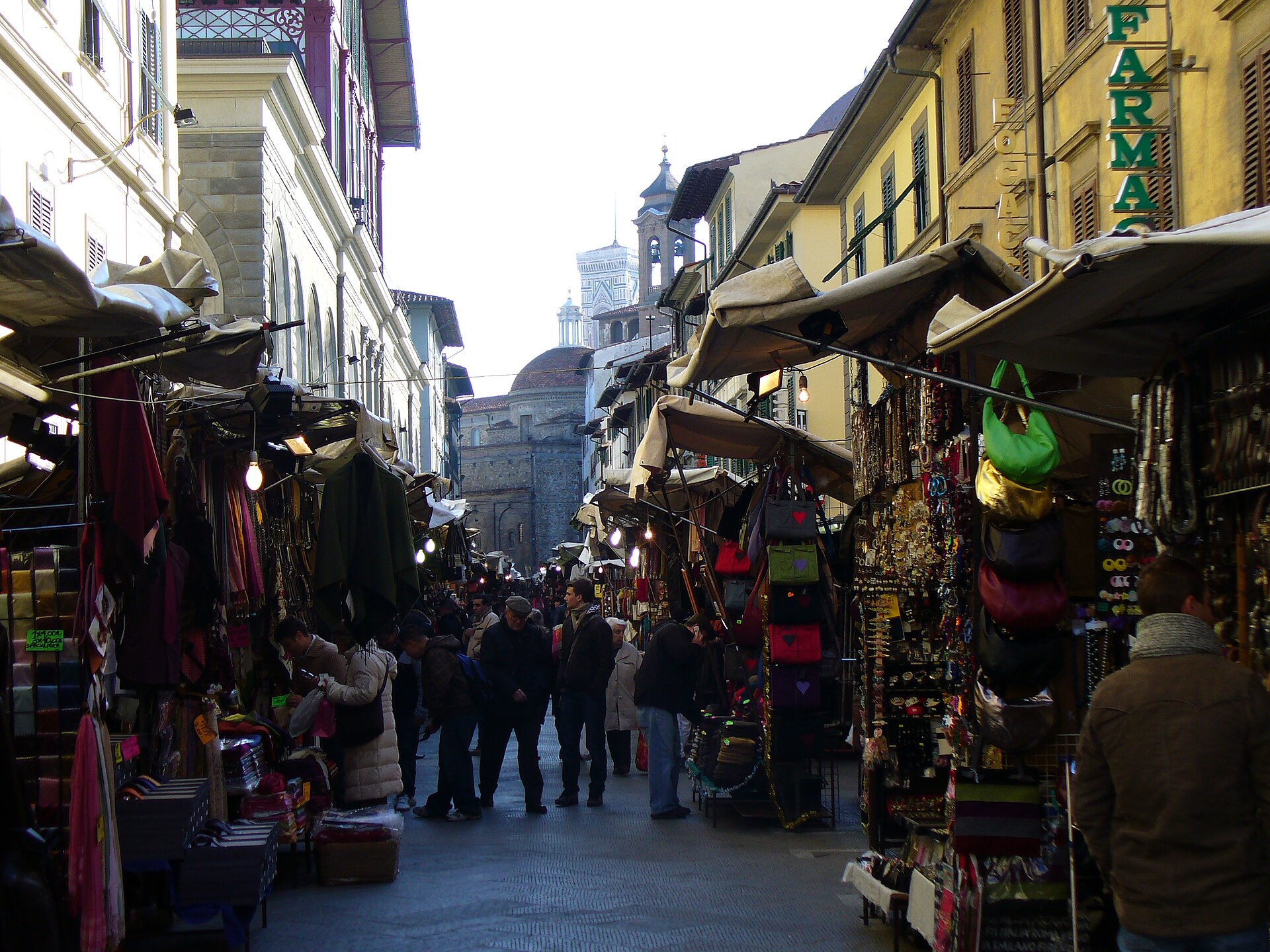 Handbags and scarves hang beside shoppers walking through Florence's San Lorenzo market toward the basilica.