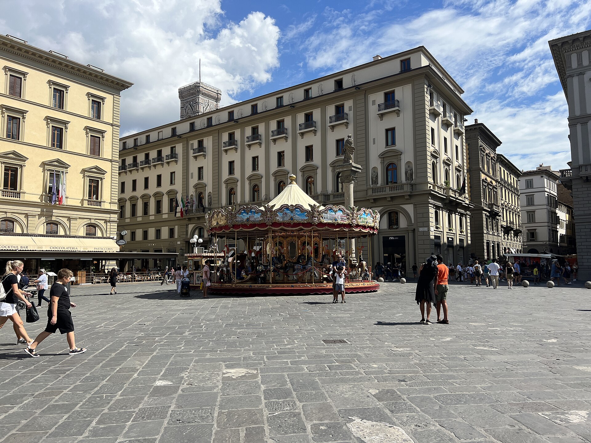 Hotel Savoy overlooks Florence’s Piazza della Repubblica behind a colorful carousel, with pedestrians crossing the stone paving.