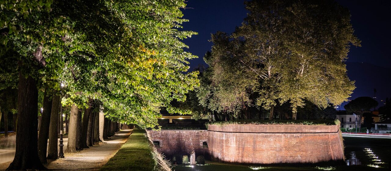 Illuminated brick ramparts and a tree-lined path on Lucca’s city walls beneath a dark evening sky.
