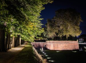 Illuminated brick ramparts and a tree-lined path on Lucca’s city walls beneath a dark evening sky.