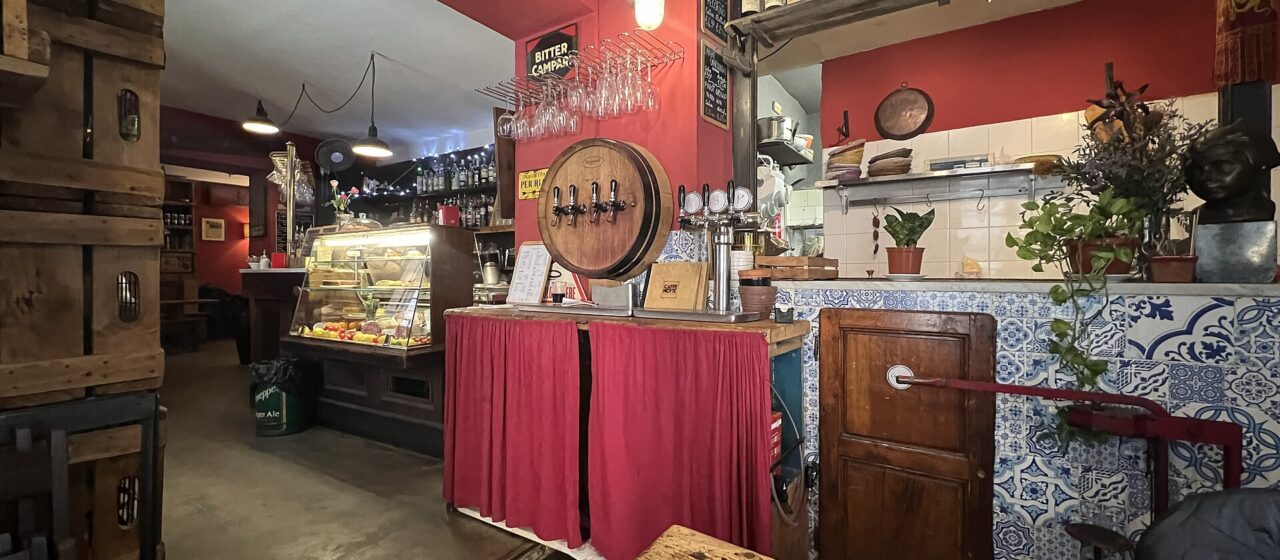 Inside a red-walled Florence wine bar with an enoteca sign, a barrel-fronted tap, bottles on shelves, a glass cabinet and tiled counter.